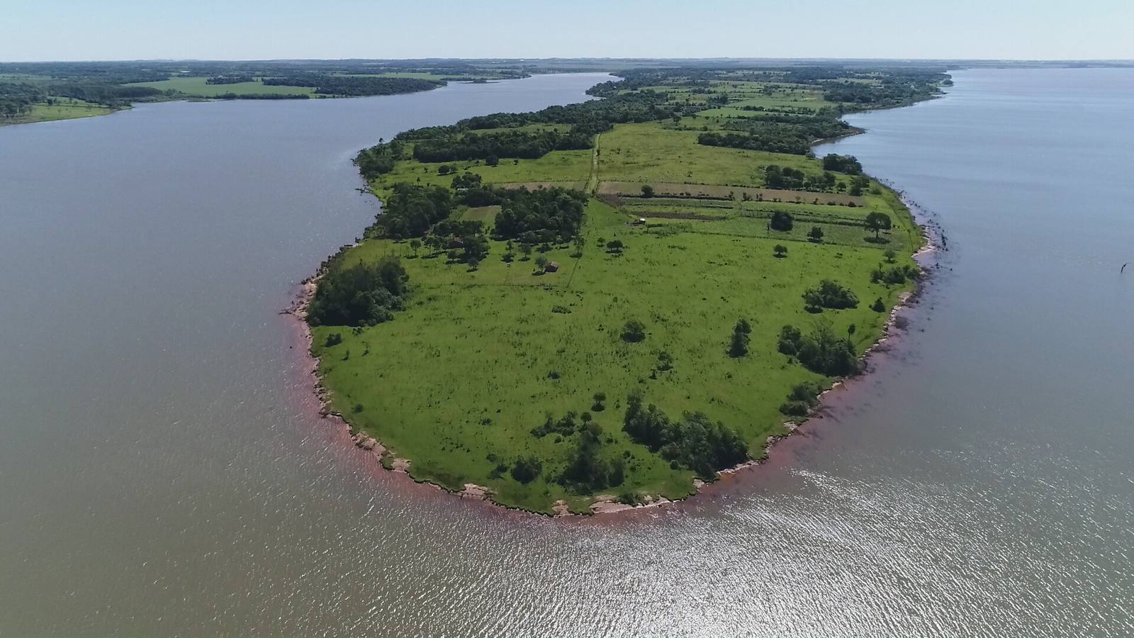 Vista aérea de la punta de la península de Zapallo: campo verde, orilla de tierra roja y agua del Lago Yguazú por tres lados.