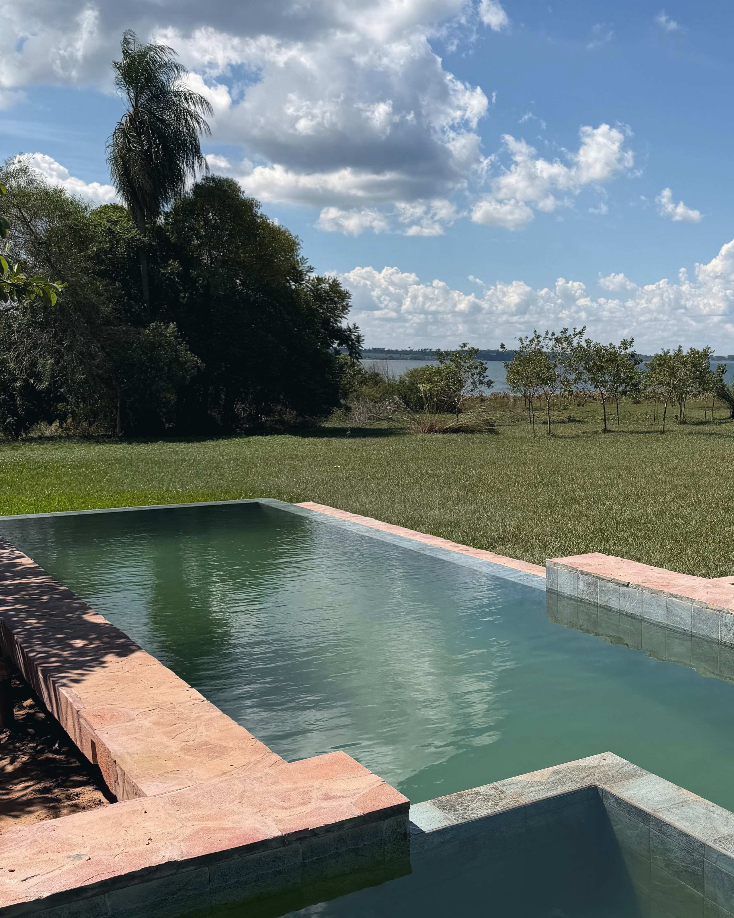 La piscina de agua verde con borde de piedra rosada y el lago entre los árboles, al fondo.